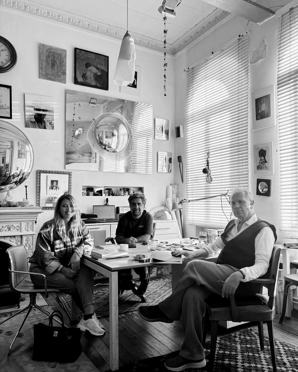 A painter’s studio crowded with framed works, where three people sit in conversation around a table.